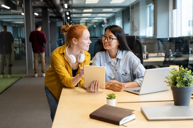 Twee jonge interculturele vrouwelijke managers die elkaar met een glimlach aankijken terwijl ze samenwerken tijdens een presentatie door de werkplek op kantoor