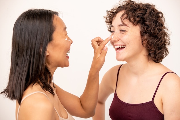 Twee jonge lachende interculturele brunettes die plezier hebben terwijl ze verjongende crème op elkaar aanbrengen