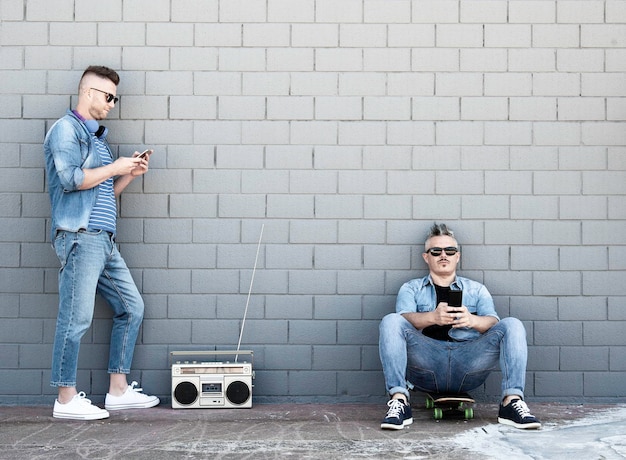 Twee jonge man kijken naar mobiele telefoon leunen tegen bakstenen muur Paar coole jongens luisteren muziek met vintage boombox stereo buiten in de straat Concept van tech sociaal netwerk en muziek trends