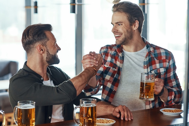 Twee jonge mannen in casual kleding handenschudden en glimlachen terwijl ze genieten van bier aan de bar in pub