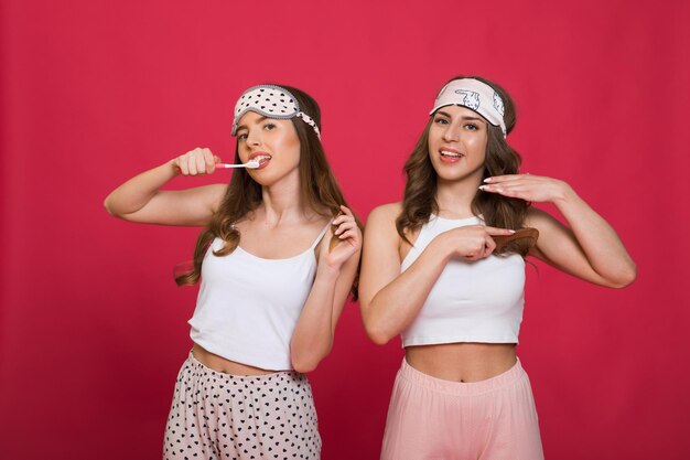 twee jonge meisjes in pyjama's en slaapmaskers dansen en zingen op een rode achtergrond met een tandenborstel