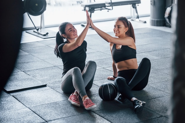 Twee jonge meisjes in sportieve kleding zitten overdag samen in de sportschool op de grond met een bal en geven high five.