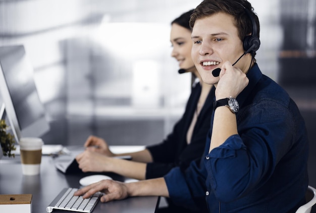 Twee jonge mensen in headsets praten met de klanten terwijl ze aan het bureau zitten in een modern kantoor Focus op man in een blauw shirt Callcenteroperators op het werk