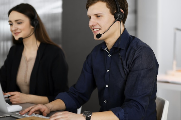 Twee jonge mensen in headsets praten met de klanten, zittend aan de balie in een modern kantoor. Focus op de mens in een blauw shirt. Callcentermedewerkers aan het werk.