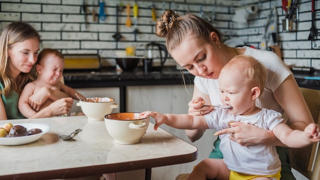 Twee jonge moeders geven samen hun gelukkige baby's melkpap in de keuken