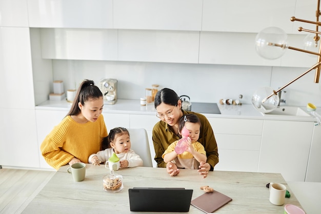 Twee jonge moeders zitten aan tafel in de keuken met hun kinderen en kijken naar tekenfilms op laptop