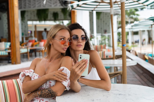 Twee jonge, mooie glimlachende hipster-vrouwen in trendy zomerzonnejurk Foto selfie maken op mobiele telefoon op terrasje Positieve modellen die plezier hebben