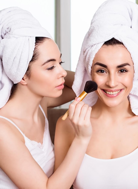 Twee jonge mooie glimlachende vrouwen met handdoeken op hun hoofd die thuis op de bank zitten te praten en een manicure en make-up doen voor het feest