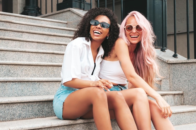 Twee jonge mooie lachende vrouw in trendy zomerkleren Sexy zorgeloze multiraciale vrouwen poseren op de straat achtergrond Positieve modellen plezier in zonnebril vrolijk en gelukkig
