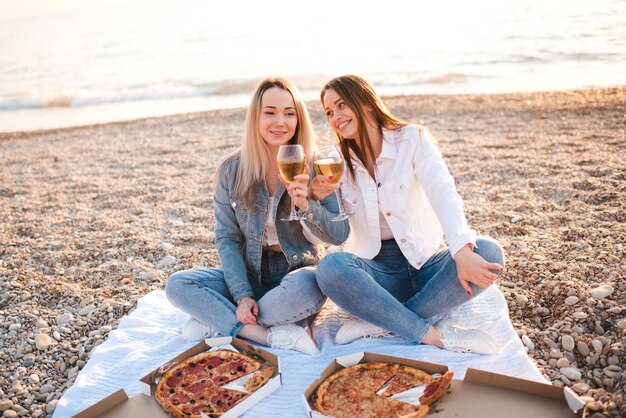 Twee jonge mooie vriendinnen die plezier hebben met pizza en wijn op het strand over de kust buiten in zonlicht. Zomer vakantie seizoen. Vriendschap. Geluk. Zusters brengen samen tijd door aan de kustlijn.