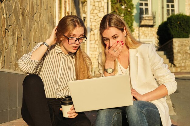 Twee jonge mooie vrouwen die plezier hebben in het kijken naar de laptop monitor zakelijke vrouwtjes zittend op trappen in de stad straat universiteitsstudenten lachen rusten