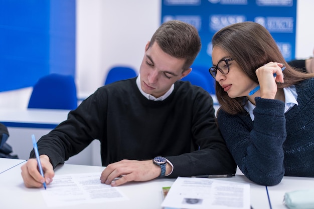 Twee jonge studenten, man en vrouw met anderen die aan een project werken in een klaslokaal dat notities schrijft en bespreekt