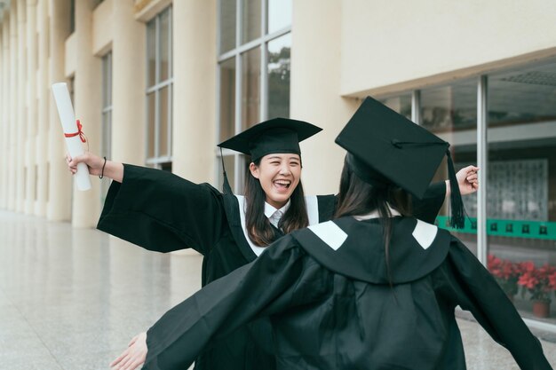 Twee jonge studentenmeisjes gekleed in zwarte afstudeerjurk glimlachend met diploma's knuffelen. vrouw met uitgestrekte armen geeft liefde aan de beste vriend op de laatste dag van de voltooide universiteit in de bouwhal.