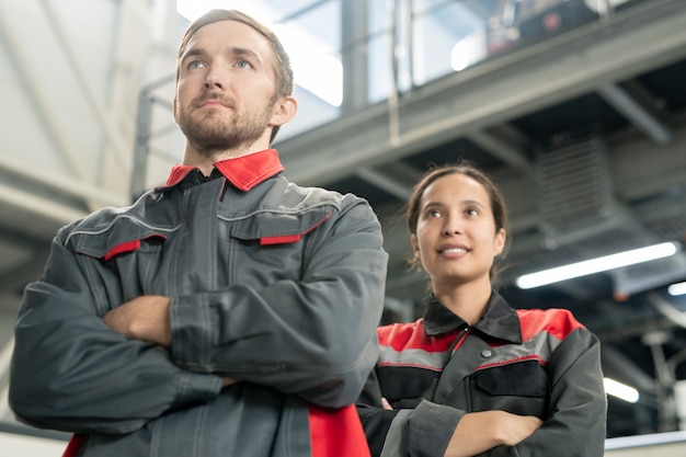 Twee jonge succesvolle arbeiders die hun armen bij de borst kruisen terwijl ze voor de camera staan in een grote moderne polymeerverwerkingsfabriek