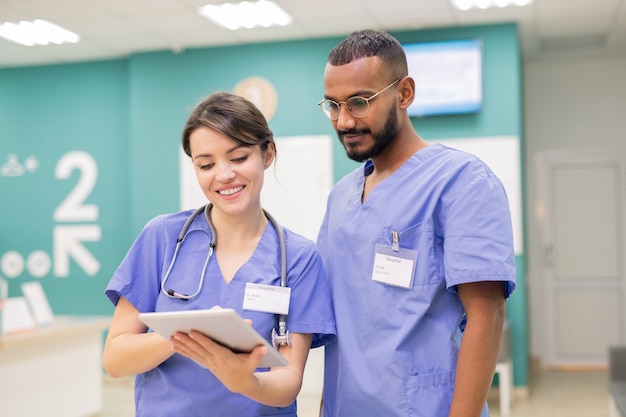 Twee jonge succesvolle clinici in uniform kijken door online medisch nieuws tijdens het scrollen in tablet op het werk