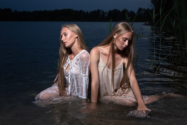 Twee jonge tweelingzusjes poseren in lichte jurken in het water van het meer op zomeravond