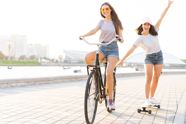 Twee jonge vrienden ontspannen op een hete zomerdag met een jonge vrouw op een fiets die haar vriend op een skateboard door een stedelijke straat sleept.