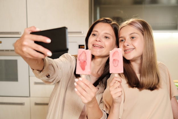 Twee jonge vrolijke vrouwtjes met lang haar selfie maken in de keuken tijdens het eten van zelfgemaakte eskimo-ijs met plakjes aardbei