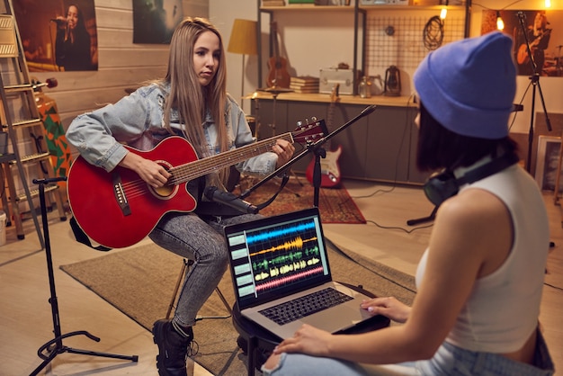 Twee jonge vrouwen die samen een nummer opnemen in de studio met behulp van laptop en gitaar spelen