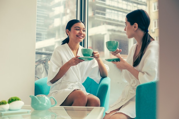 Twee jonge vrouwen in badjassen hebben een interessant gesprek terwijl ze in fauteuils zitten en thee drinken