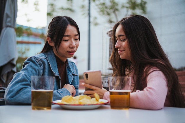 Twee jonge vrouwen kijken naar foto's op een smartphone terwijl ze op het terras van de bar zitten