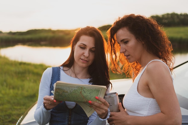 Twee jonge vrouwen reizen op de wegen met een auto die is gestopt aan de kant van de weg en kijken naar de kaart die koffie drinkt uit een thermoskan Vakantieconcept