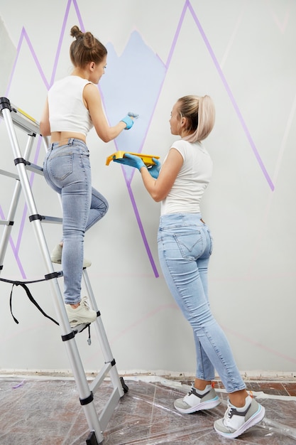 Twee jonge vrouwen schilderen muur met verfroller en gebruiken plakband terwijl ze op een ladder staan