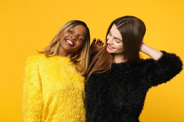 Twee jonge vrouwen vrienden Europese en Afrikaanse Amerikaan in zwarte gele kleding staande poseren geïsoleerd op fel oranje muur achtergrond, studio portret. Mensen levensstijl concept. Bespotten kopie ruimte.