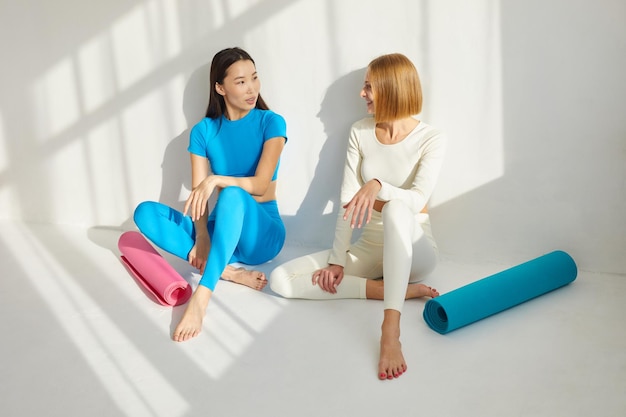 Foto twee jonge vrouwen zitten in een yoga les en glimlachen en praten.