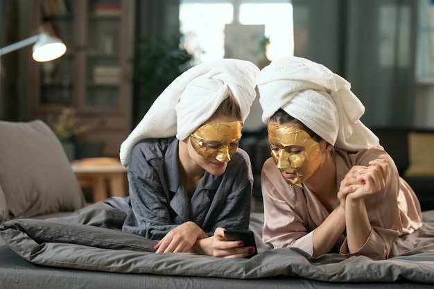 Twee jonge vrouwtjes in zijden pyjama's, handdoeken op hoofden en gouden cosmetische maskers op gezichten die ontspannen op bed en scrollen in smartphone