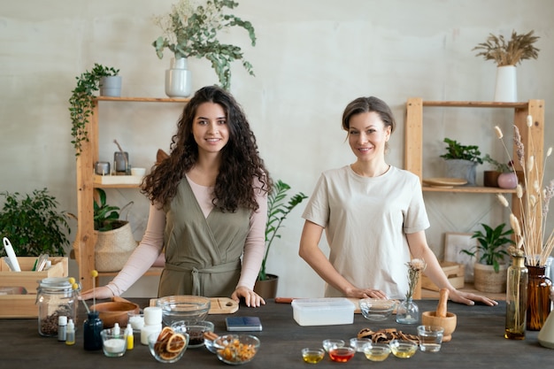 Twee jonge vrouwtjes staan bij de tafel terwijl een van hen harde zeepmassa in een plastic bak snijdt voordat ze natuurlijke cosmetische producten maakt