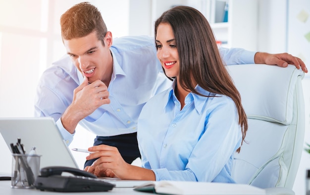 Twee jonge zakenmensen praten op kantoor. vrouw zit aan het bureau en typt op laptop, achter haar staat man en kijkt naar laptop.