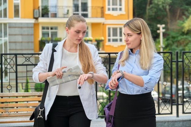 Twee jonge zakenvrouwen die buiten lopen en praten, stadsachtergrond, vrouwelijke kantoormedewerkers, vrouw kijken naar polshorloge