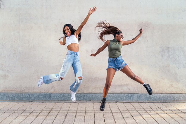 Twee jonge zwarte meisjes springen blij met een grijze betonnen muur op de achtergrond, concept van vriendschap en stedelijke levensstijl