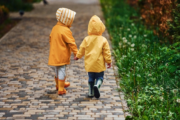 Twee kinderen in gele waterdichte mantels en laarzen houden elkaars handen vast en maken samen een wandeling buiten na de regen