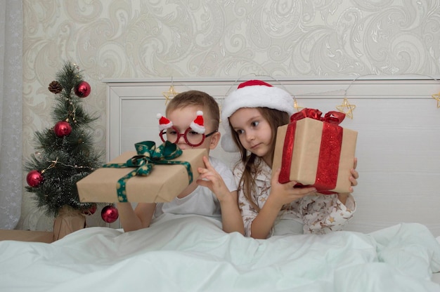 Twee kinderen in pyjama zittend op het bed op kerstavond. Broer en zus openen nieuwjaarsgeschenken.