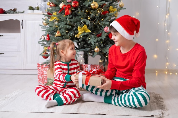 twee kinderen jongen en meisje in kerstpyjama met geschenkdozen die thuis onder de kerstboom zitten