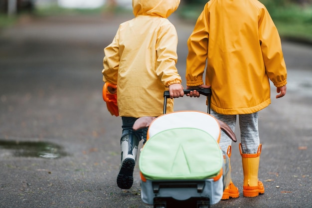 Twee kinderen met paraplukoffer en gele waterdichte mantels en laarzen die samen buiten lopen na de regen