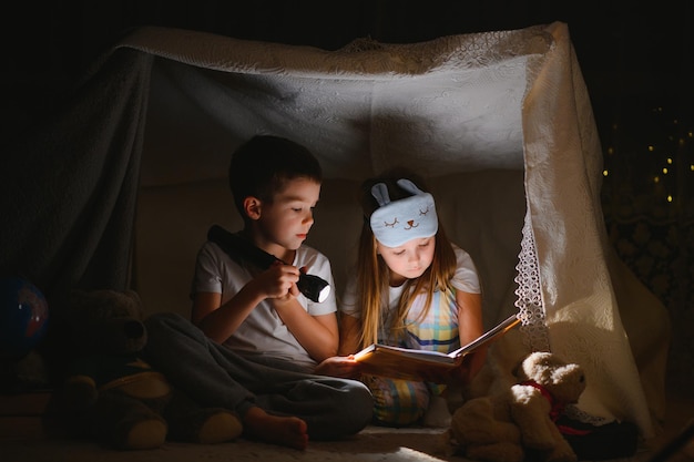 Twee kinderen met zaklamp lezen een boek onder een deken als tent