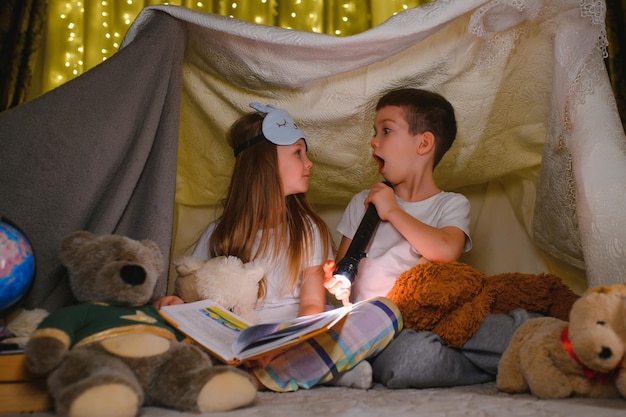 Twee kinderen met zaklamp lezen een boek onder een deken als tent