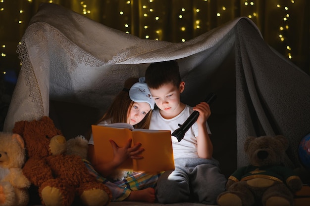 Twee kinderen met zaklamp lezen een boek onder een deken als tent