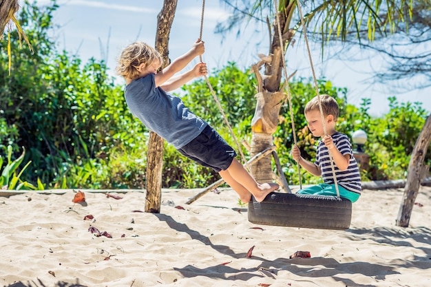 Twee kleine blonde jongens die plezier hebben op de schommel aan de tropische zandkust