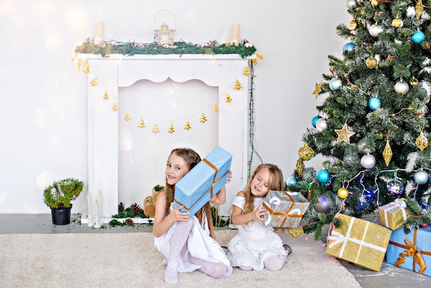 Twee kleine blonde meisjes in witte jurken spelen bij de open haard en de kerstboom met dozen.