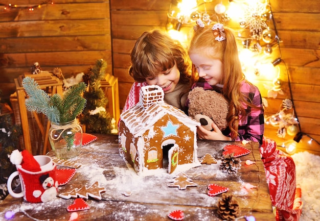 Twee kleine kinderen - een jongen met krullend haar en een roodharig meisje bereiden een peperkoekhuis voor en hebben plezier tegen de achtergrond van kerstverlichting en decor. kersttraditie