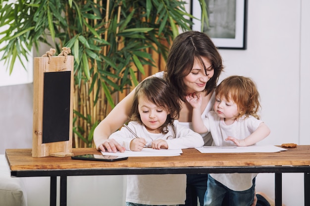 Twee kleine kinderen meisjes schattig mooi en blij met moeder thuis aan tafel zitten en tekenen