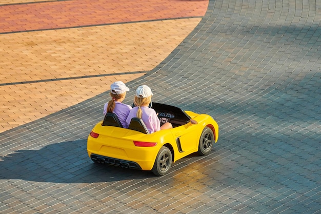 Twee kleine meisjes gele speelgoedauto rijden op de stoep in pretpark. Kinderen rijden elektrische auto met afstandsbediening. Batterijvoertuig voor kinderentertainment