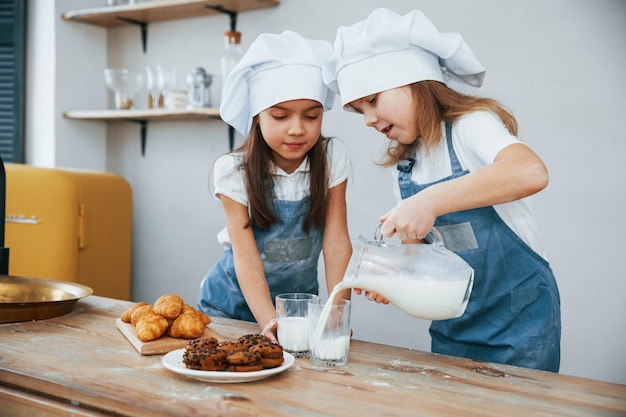 Twee kleine meisjes in blauwe chef-kok uniform gieten melk in glazen op de keuken met koekjes op tafel