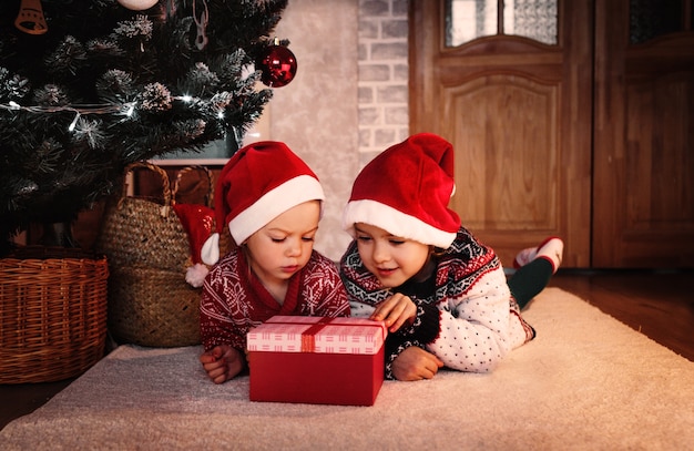 Twee kleine meisjes in een kerstmuts openen een kerstcadeau doos onder de boom