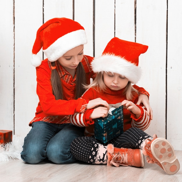 Twee kleine meisjes in rode hoeden zittend op de vloer met geschenken in de buurt van de nieuwe jaarboom