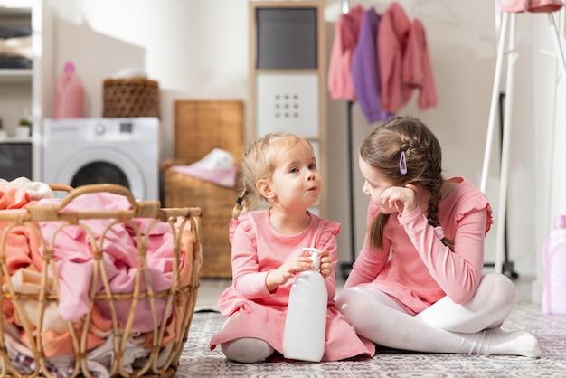 Twee kleine meisjes in roze jurken praten zittend op de vloer van de wasruimte bij een mand met kleren, het jongere kind houdt een fles vloeistof vast